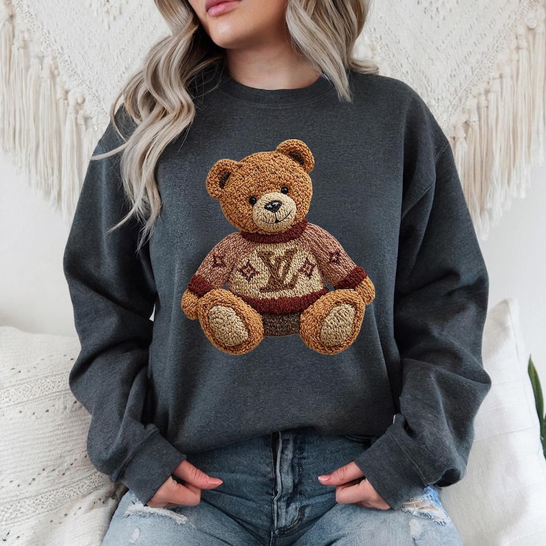 May include: Dark gray sweatshirt featuring a detailed teddy bear design. The bear is brown with a sweater that has a logo in the center. The sweatshirt is a solid color and has long sleeves.