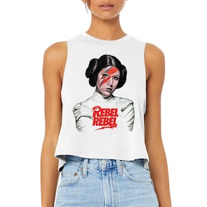 May include: White sleeveless crop top featuring a graphic of a woman with a lightning bolt on her face and the words "Rebel Rebel" in red. The image is a fusion of pop culture icons.