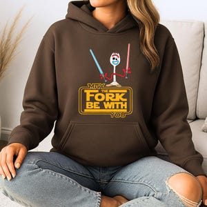Op de afbeelding: Een donkerbruine hoodie met een afbeelding van een wit spork-personage dat een blauw en een rood lichtzwaard vasthoudt. Onder het personage staat de gele tekst: MAY THE FORK BE WITH YOU. De hoodie heeft lange mouwen en een kangoeroezak aan de voorzijde.