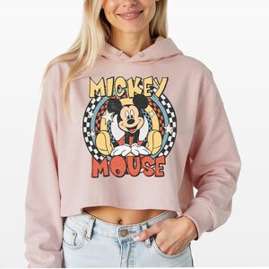 May include: A light pink cropped hoodie featuring a graphic print of Mickey Mouse. The design includes the text "MICKEY MOUSE" in yellow and red, surrounded by a black and white checkered circle, blue rays, and white stars on a distressed background.