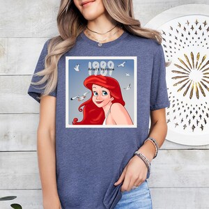 May include: A blue t-shirt with a graphic of Ariel from The Little Mermaid, with the text "1989 Ariel's Version." The graphic shows Ariel with red hair and seagulls. The shirt is a casual wear item.