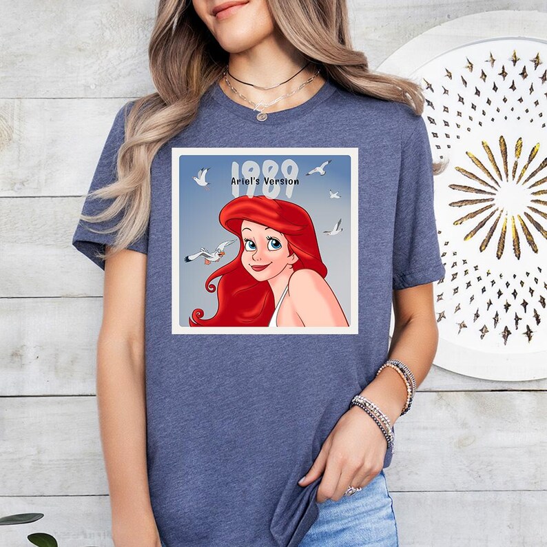 May include: A blue t-shirt with a graphic of Ariel from The Little Mermaid, with the text "1989 Ariel's Version." The graphic shows Ariel with red hair and seagulls. The shirt is a casual wear item.
