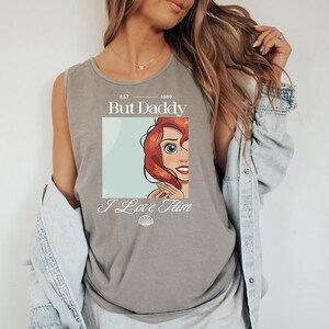 May include: Gray tank top featuring a graphic of a cartoon character with red hair and the text "But Daddy I Love Him" with a seashell graphic. The top has a crew neck and is sleeveless.