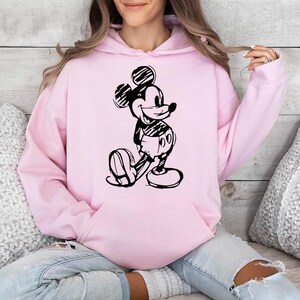 May include: A light pink hoodie featuring a black and white sketch of Mickey Mouse. The hoodie has a kangaroo pocket and a drawstring hood. The design is a classic cartoon character.