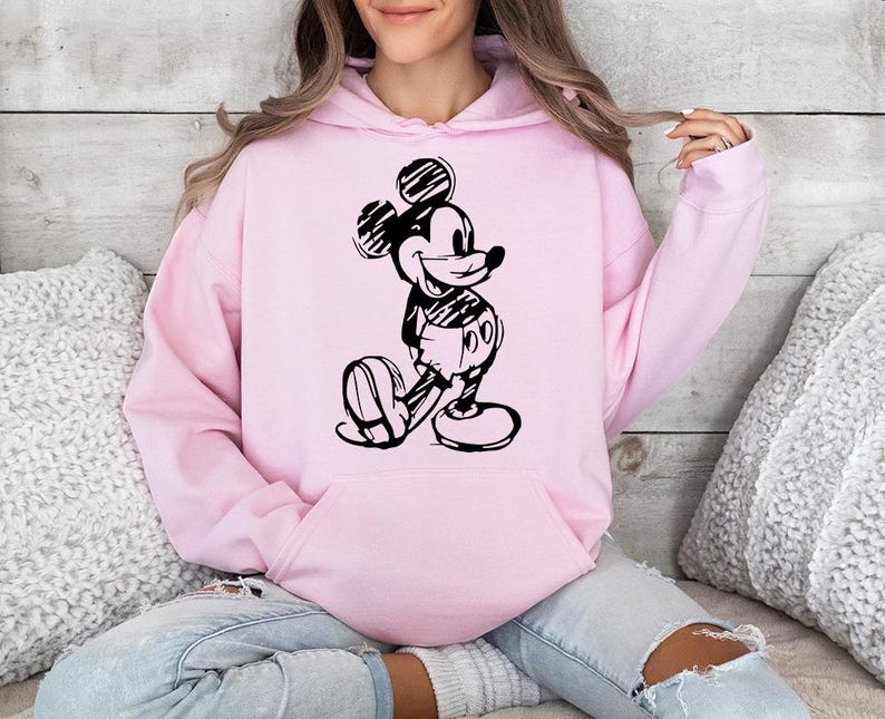 May include: A light pink hoodie featuring a black and white sketch of Mickey Mouse. The hoodie has a kangaroo pocket and a drawstring hood. The design is a classic cartoon character.