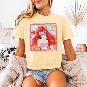 K&ouml;nnte beinhalten: Hellgelbes T-Shirt mit einem Aufdruck einer rothaarigen Meerjungfrau mit herzf&ouml;rmiger Sonnenbrille, die eine Blase bl&auml;st. Die Grafik ist von Muscheln und Seesternen umgeben. Das Shirt wird mit Jeansshorts getragen.