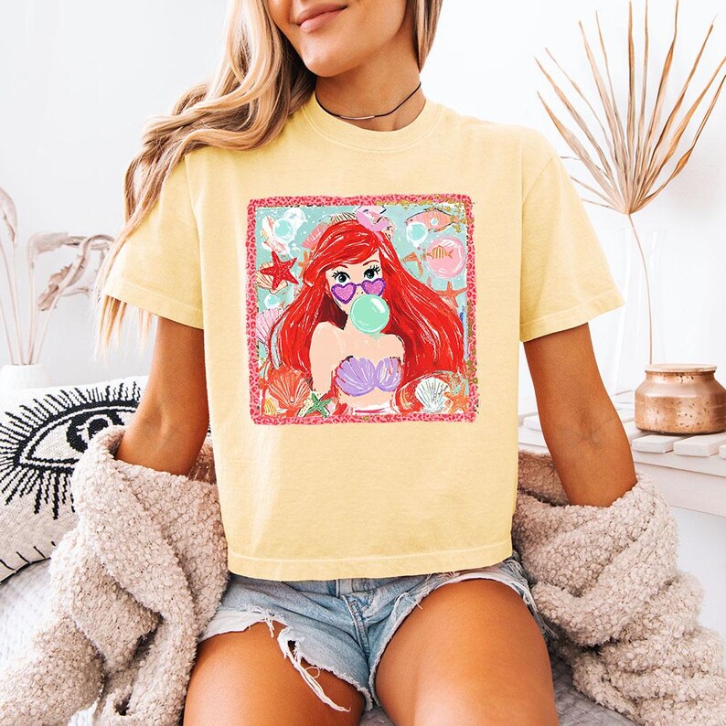 K&ouml;nnte beinhalten: Hellgelbes T-Shirt mit einem Aufdruck einer rothaarigen Meerjungfrau mit herzf&ouml;rmiger Sonnenbrille, die eine Blase bl&auml;st. Die Grafik ist von Muscheln und Seesternen umgeben. Das Shirt wird mit Jeansshorts getragen.