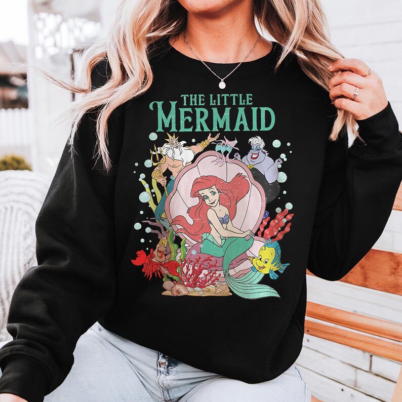 May include: A black long-sleeve sweatshirt featuring a graphic print of The Little Mermaid characters including Ariel, King Triton, Ursula, Sebastian, and Flounder. The design includes the text THE LITTLE MERMAID in teal letters above the characters.