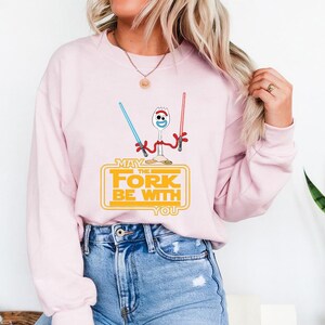May include: A light pink crewneck sweatshirt featuring a graphic of a white spork character with googly eyes holding a blue and a red lightsaber. Below the character, the yellow text reads: MAY THE FORK BE WITH YOU in a stylized rectangular frame.