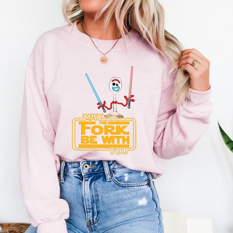 May include: A light pink crewneck sweatshirt featuring a graphic of a white spork character with googly eyes holding a blue and a red lightsaber. Below the character, the yellow text reads: MAY THE FORK BE WITH YOU in a stylized rectangular frame.