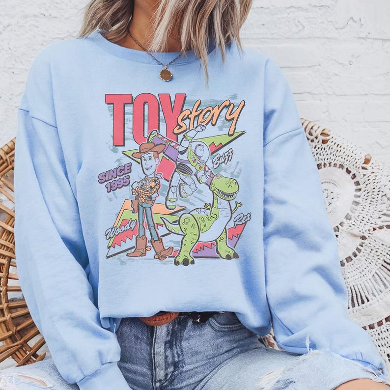May include: A light blue crewneck sweatshirt featuring a graphic print of Toy Story characters Woody, Buzz Lightyear, and Rex. The design includes the text TOY story, SINCE 1995, Woody, Buzz, and Rex, set against a colorful geometric background.