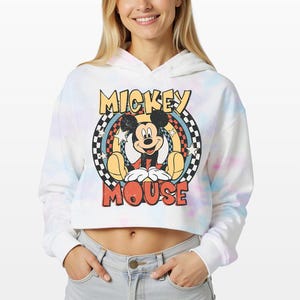 May include: A white cropped hoodie with a pastel tie-dye pattern featuring a graphic of Mickey Mouse. The design includes a checkered circular border, yellow stars, and the text MICKEY MOUSE in a distressed orange and yellow font style.