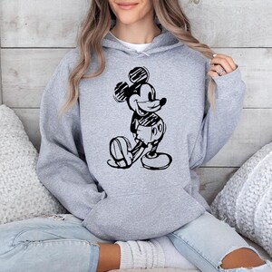 May include: A light gray hooded sweatshirt featuring a black and white sketch of Mickey Mouse. The cartoon character is depicted in a classic pose, with his hands behind his back. The sweatshirt has a drawstring hood and a kangaroo pocket.