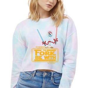 May include: A long-sleeve crop sweatshirt with a pastel tie-dye pattern in blue, pink, and white. The front features a graphic of a white plastic spork character holding a blue and red lightsaber, with the yellow text May The Fork Be With You.