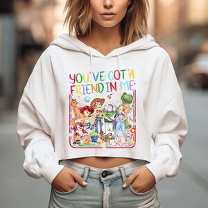 May include: A white cropped hoodie featuring a colorful graphic print of various animated toy characters. The text "YOU'VE GOT A FRIEND IN ME" is printed in multi-colored letters above the characters, all enclosed within a thin red and blue border.