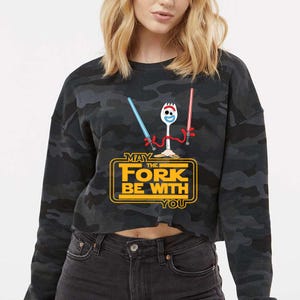 May include: A black and grey camouflage print long-sleeve sweatshirt featuring a graphic of a white spork character holding a blue and a red lightsaber. Below the character, the yellow text reads MAY THE FORK BE WITH YOU in a bold font.