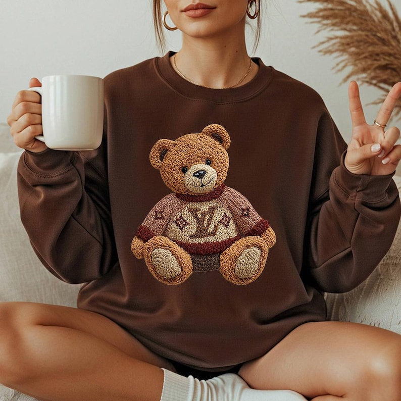 May include: A brown sweatshirt featuring a detailed embroidered teddy bear wearing a sweater with a logo. The bear has a light brown fur and is wearing a sweater with a burgundy and light brown design. The sweatshirt is a solid brown color.