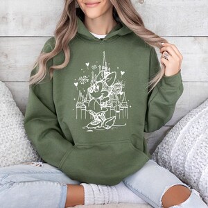 May include: Olive green hooded sweatshirt featuring a white graphic of a cartoon character in front of a castle with fireworks. The sweatshirt has a front pocket and a drawstring hood. The design is a whimsical illustration.