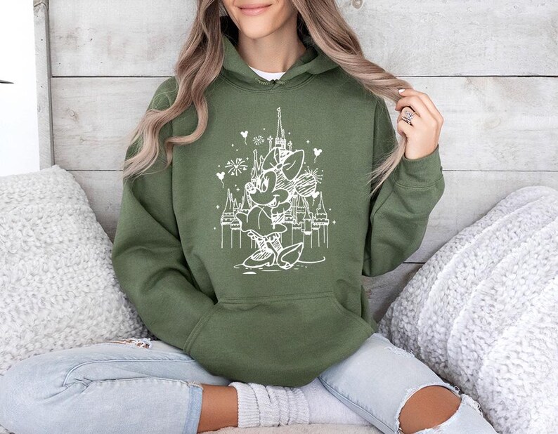 May include: Olive green hooded sweatshirt featuring a white graphic of a cartoon character in front of a castle with fireworks. The sweatshirt has a front pocket and a drawstring hood. The design is a whimsical illustration.
