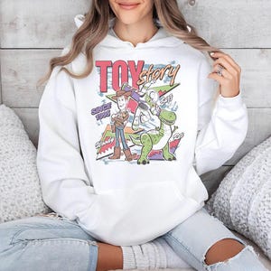 May include: A white hooded sweatshirt featuring a colorful graphic print of the Toy Story characters Woody, Buzz Lightyear, and Rex. The design includes the text Toy Story, Since 1995, Woody, Buzz, and Rex, set against a geometric background pattern.