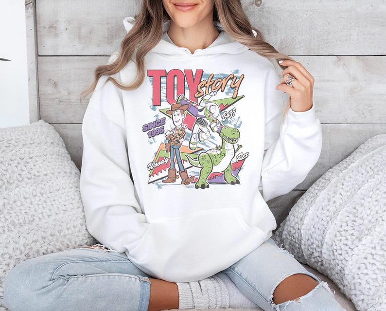 May include: A white hooded sweatshirt featuring a colorful graphic print of the Toy Story characters Woody, Buzz Lightyear, and Rex. The design includes the text Toy Story, Since 1995, Woody, Buzz, and Rex, set against a geometric background pattern.