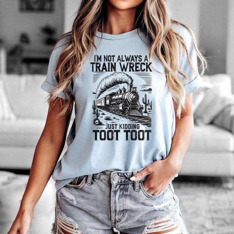 May include: Light blue t-shirt with a black and white train graphic. The text on the shirt reads "I'M NOT ALWAYS A TRAIN WRECK JUST KIDDING TOOT TOOT". The graphic features a steam train on tracks with cacti.