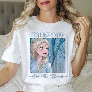 May include: White t-shirt with a blue and white illustration of a woman with long blonde hair. The text "It's Like Snow On The Beach" is printed on the shirt.