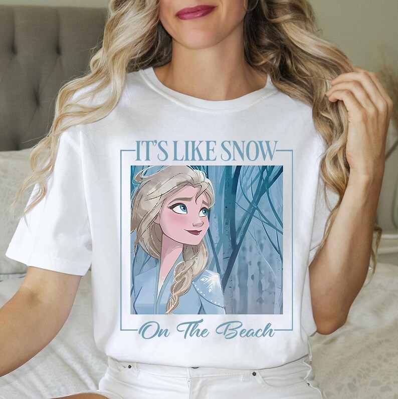 May include: White t-shirt with a blue and white illustration of a woman with long blonde hair. The text "It's Like Snow On The Beach" is printed on the shirt.