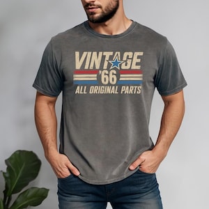 Puede incluir: Camiseta gris con el texto "VINTAGE '66 ALL ORIGINAL PARTS" en una fuente desgastada. El dise&ntilde;o incluye una estrella azul y rayas rojas y azules, d&aacute;ndole un aire retro. La camiseta es de cuello redondo.