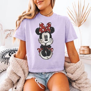 Puede incluir: Una camiseta de manga corta de color morado claro con un estampado gr&aacute;fico desgastado de Minnie Mouse. El personaje aparece con las manos bajo la barbilla, con un lazo rojo y blanco de lunares y sentada sobre una falda de lunares rojos y blancos.