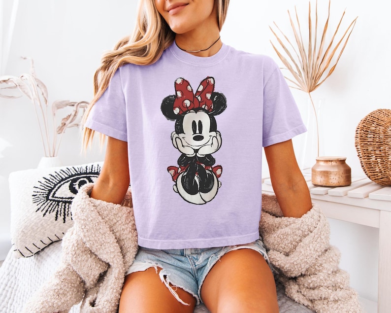 Puede incluir: Una camiseta de manga corta de color morado claro con un estampado gr&aacute;fico desgastado de Minnie Mouse. El personaje aparece con las manos bajo la barbilla, con un lazo rojo y blanco de lunares y sentada sobre una falda de lunares rojos y blancos.