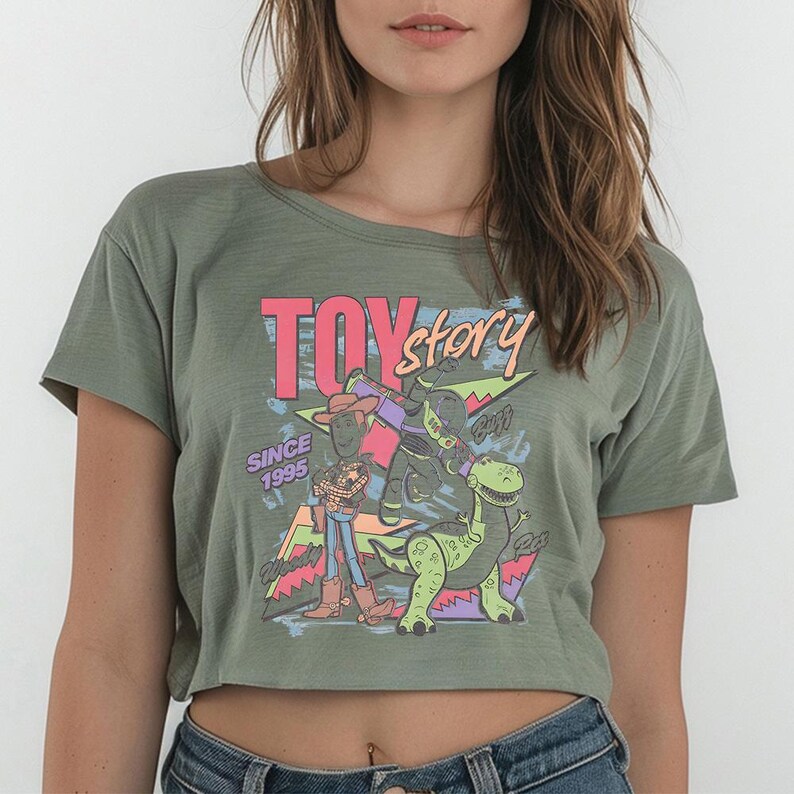 K&ouml;nnte beinhalten: Ein olivgr&uuml;nes kurz&auml;rmeliges bauchfreies Oberteil mit einem Grafikdruck von Woody, Buzz Lightyear und Rex aus Toy Story. Das Design enth&auml;lt den Text "TOY story" in Pink und Orange, "SINCE 1995" in Lila sowie bunte geometrische Formen im Hintergrund.