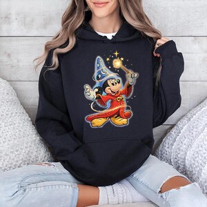 May include: A navy blue hooded sweatshirt featuring a colorful graphic of Mickey Mouse dressed as a wizard, holding a wand with sparkling stars. The cartoon character is wearing a blue wizard hat and a red robe. The sweatshirt has a drawstring hood.