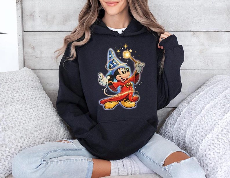 May include: A navy blue hooded sweatshirt featuring a colorful graphic of Mickey Mouse dressed as a wizard, holding a wand with sparkling stars. The cartoon character is wearing a blue wizard hat and a red robe. The sweatshirt has a drawstring hood.