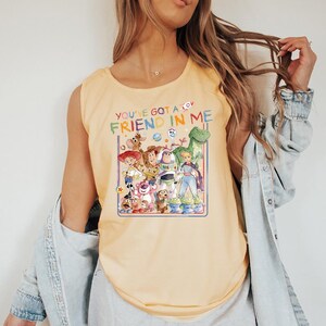 Pu&ograve; includere: Una canotta giallo chiaro con una grafica colorata dei personaggi del film Toy Story. Il testo "YOU'VE GOT A FRIEND IN ME" &egrave; stampato sopra i personaggi in un font arcobaleno.