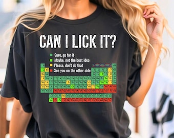Op de afbeelding: Zwart T-shirt met de tekst "CAN I LICK IT?" en een periodiek systeem der elementen. Het ontwerp bevat een kleurgecodeerde sleutel die de veiligheid van het likken van elk element aangeeft. Een humoristisch, wetenschappelijk T-shirt.