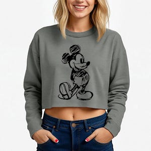 May include: A gray cropped sweatshirt featuring a black sketch of Mickey Mouse. The long-sleeved top has a crew neck and is paired with blue jeans. The Mickey Mouse graphic is centered on the front of the shirt.