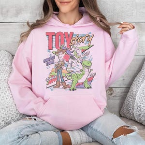 May include: A light pink hooded sweatshirt featuring a graphic print of Toy Story characters Woody, Buzz Lightyear, and Rex. The design includes the text "TOY story", "SINCE 1995", "Woody", "Buzz", and "Rex" with colorful geometric shapes in the background.