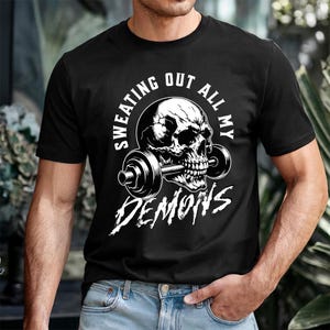 Może przedstawiać: Czarna koszulka z białym nadrukiem czaszki trzymającej sztangę. Tekst "SWEATING OUT ALL MY DEMONS" jest wydrukowany wokół czaszki i pod nią. Koszulka wykonana jest z miękkiego materiału.