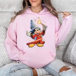 May include: A light pink hoodie featuring a graphic of Mickey Mouse dressed as a wizard, holding a wand with sparkling stars. The cartoon character is wearing a blue wizard hat and a red robe. The hoodie has a front pocket and a drawstring hood.
