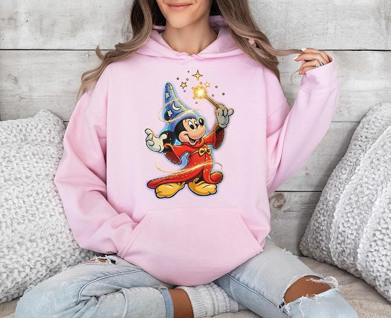 May include: A light pink hoodie featuring a graphic of Mickey Mouse dressed as a wizard, holding a wand with sparkling stars. The cartoon character is wearing a blue wizard hat and a red robe. The hoodie has a front pocket and a drawstring hood.