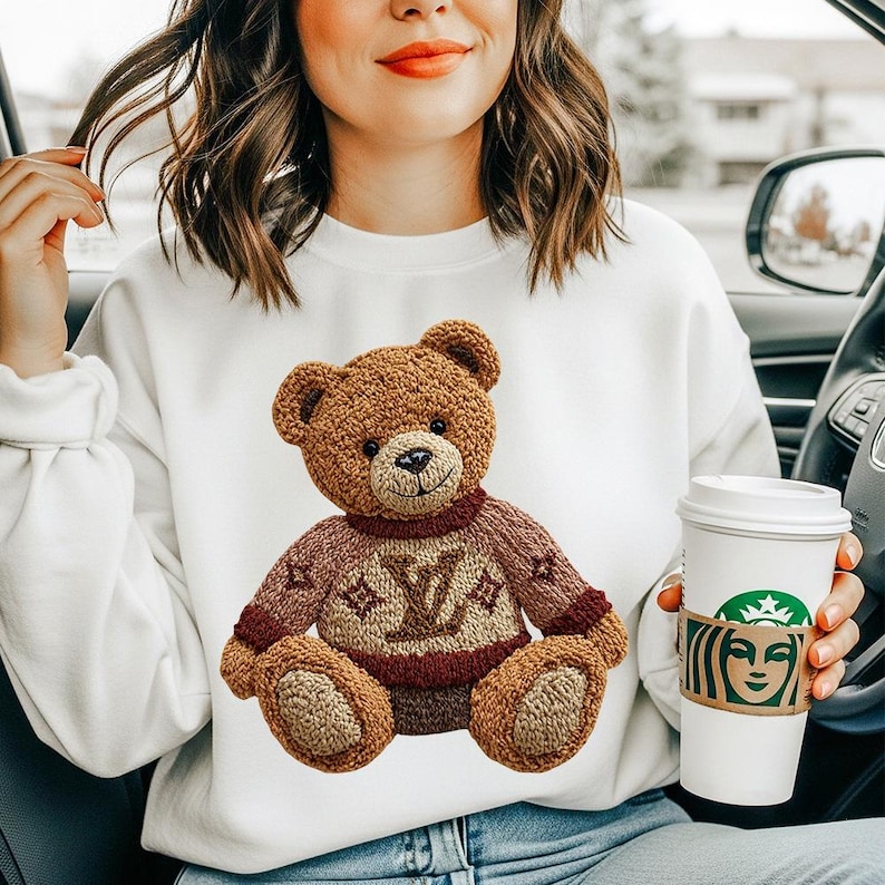 May include: White sweatshirt featuring a brown teddy bear design. The teddy bear has a brown body, a red sweater with a logo, and a smiling face. The sweatshirt is a comfortable, casual style.