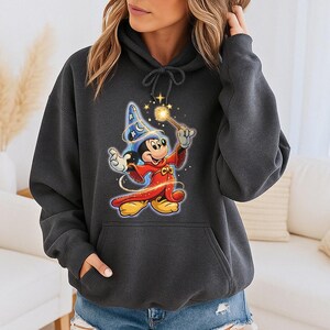 May include: Dark gray hoodie featuring a colorful graphic of Mickey Mouse as a sorcerer, holding a glowing wand. The design includes a blue wizard hat and a red robe. The hoodie has a drawstring hood and a front pocket.