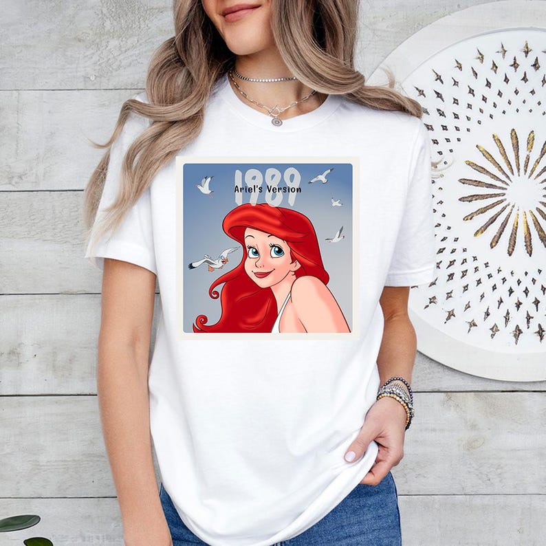 May include: White t-shirt with a square graphic of Ariel from The Little Mermaid. The graphic shows Ariel, seagulls, and the text "1989 Ariel's Version" against a blue backdrop. The shirt has a crew neck.