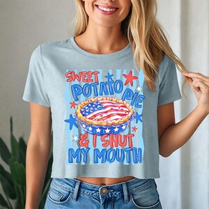 Op de afbeelding: Een lichtblauw T-shirt met korte mouwen en een afbeelding van een taart met een Amerikaans vlagpatroon. De tekst "SWEET POTATO PIE & I SHUT MY MOUTH" staat in rode en blauwe letters, omringd door rode en blauwe sterren en verticale strepen.