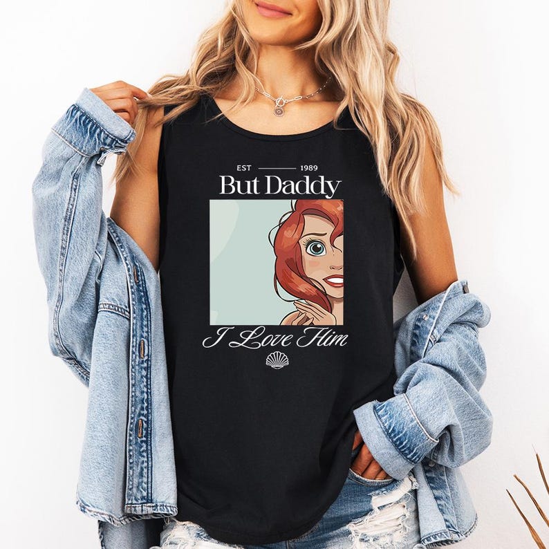 May include: Black tank top featuring a graphic of a cartoon character with red hair and the text "But Daddy I Love Him" with a seashell graphic. The tank top has the text "EST 1989" above the main text.
