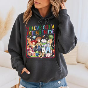 May include: A charcoal gray hooded sweatshirt featuring a colorful graphic print of various animated characters from the Toy Story franchise. The text "YOU'VE GOT A FRIEND IN ME" is printed in multicolored letters above the character group illustration.