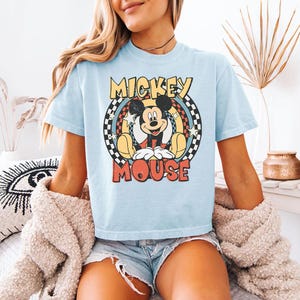 Pu&ograve; includere: Una t-shirt azzurro chiaro con una grafica di Mickey Mouse centrata all'interno di un design circolare. La grafica include il testo MICKEY MOUSE in giallo e rosso, circondato da un bordo a scacchi bianco e nero, stelle gialle e cerchi.