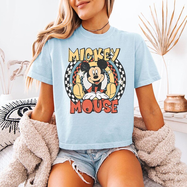 Pu&ograve; includere: Una t-shirt azzurro chiaro con una grafica di Mickey Mouse centrata all'interno di un design circolare. La grafica include il testo MICKEY MOUSE in giallo e rosso, circondato da un bordo a scacchi bianco e nero, stelle gialle e cerchi.