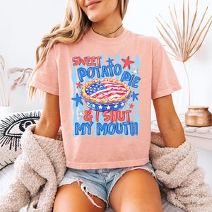 Op de afbeelding: Een perzikkleurig t-shirt met een afbeelding van een taart met een Amerikaans vlagpatroon. De tekst "SWEET POTATO PIE & I SHUT MY MOUTH" staat in blauwe en rode letters, omringd door blauwe verticale strepen en rode en blauwe sterren.